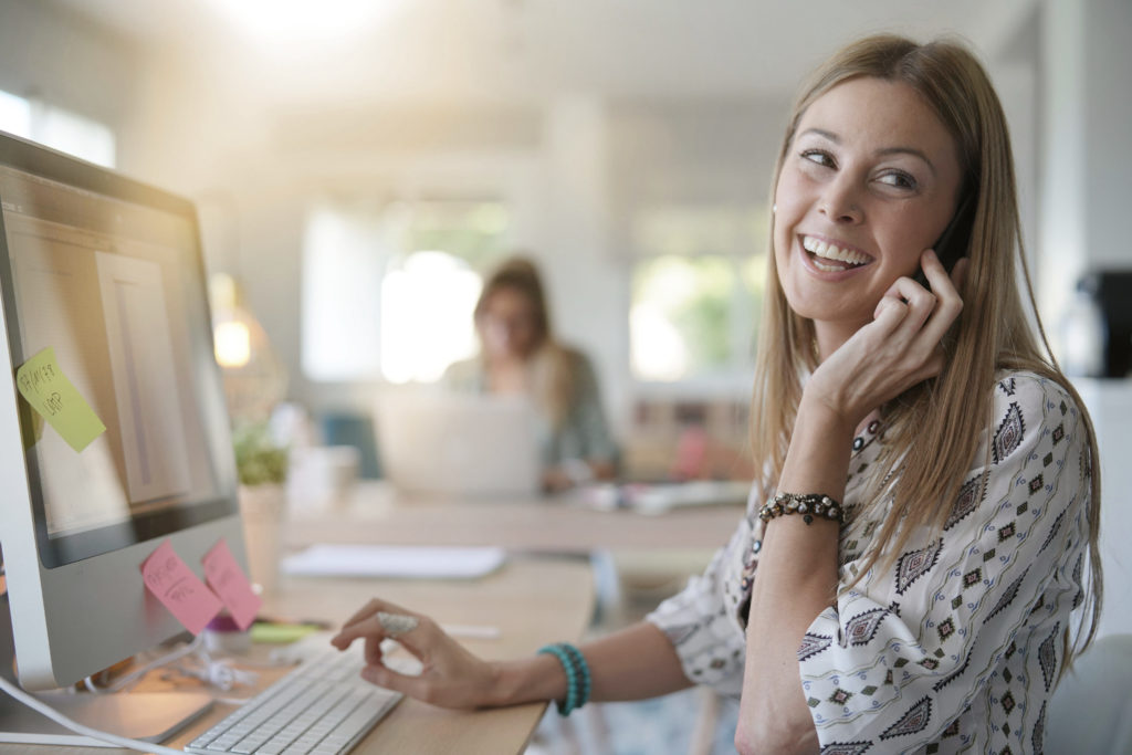 Jeune femme souriante au téléphone, travaillant sur un ordinateur dans un bureau lumineux, illustrant le premier contact pour un bilan de compétences et une reconversion professionnelle.